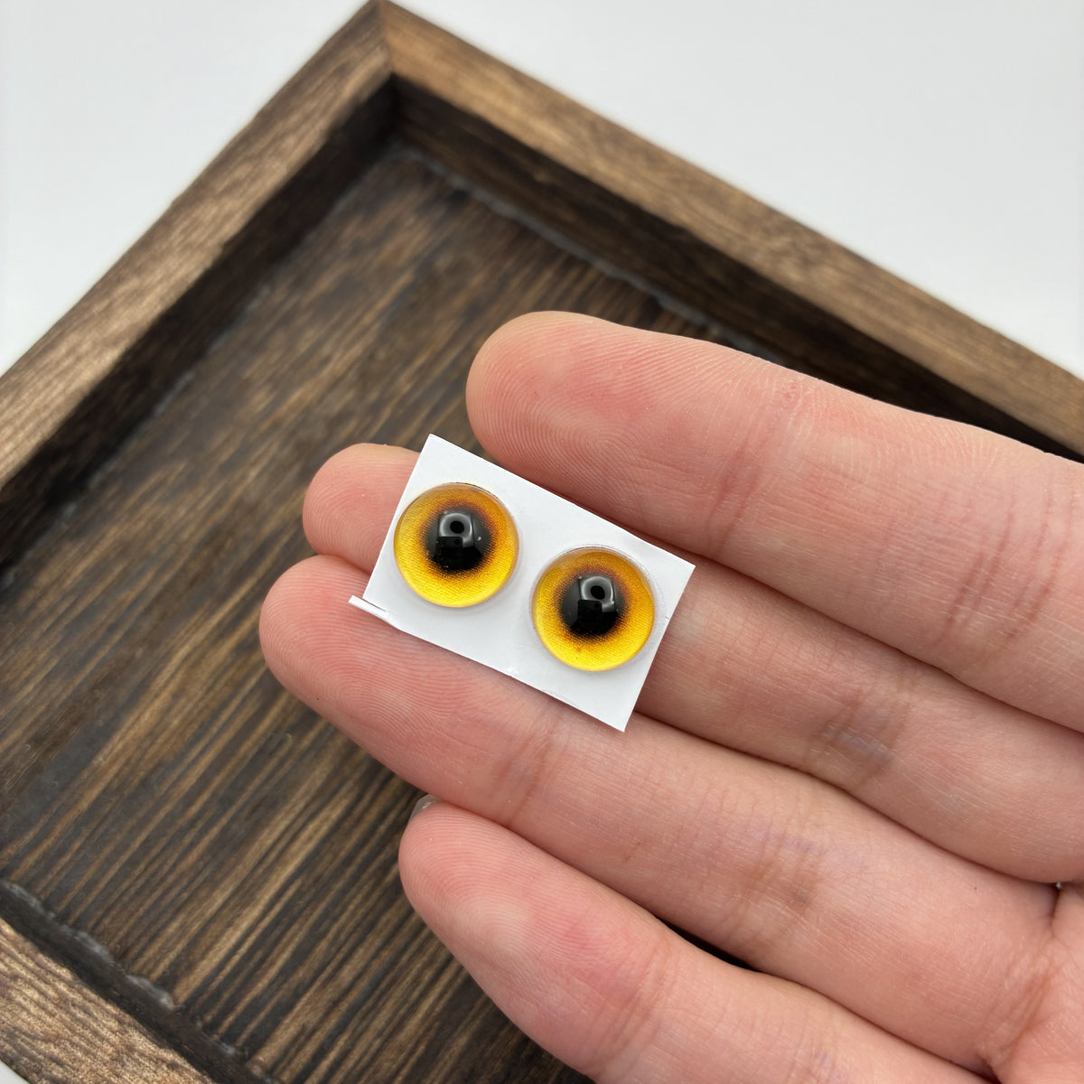 Paire d'yeux en verre faits main 10 mm ambre sur support papier, tenue en main pour montrer la taille