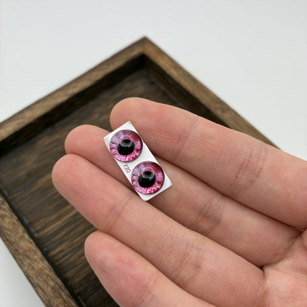 Paire d'yeux en verre faits main 10 mm rose sur support papier, tenue en main pour montrer la taille